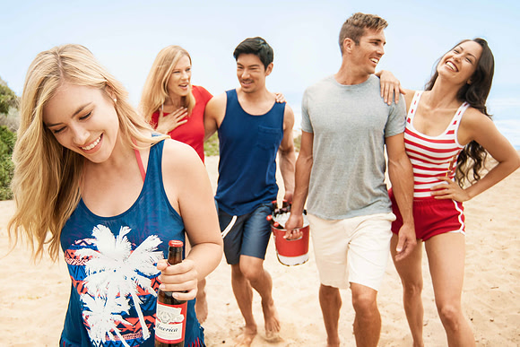 Lifestyle advertising photograph of four friends walking together on the beach while carrying bottled drinks.