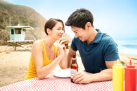 Advertising photograph of a couple smiling over food and bottled drinks at a beach picnic table.