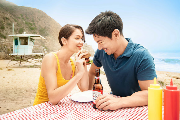 Advertising photograph of a couple smiling over food and bottled drinks at a beach picnic table.