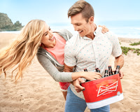 Lifestyle advertising photograph of a smiling couple carrying a red beverage cooler across a sandy beach.