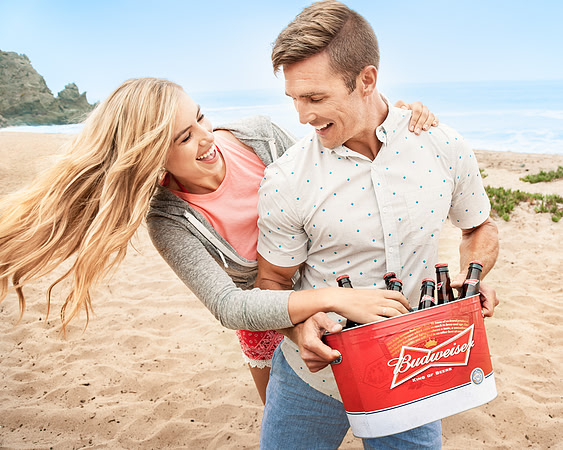 Lifestyle advertising photograph of a smiling couple carrying a red beverage cooler across a sandy beach.