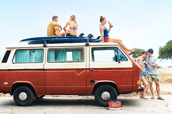 Wide lifestyle photograph of friends sitting on and around a vintage van during a beach outing.