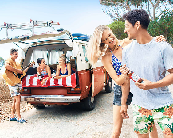 Lifestyle advertising photograph of a smiling couple holding a bottled drink beside a vintage beach van.
