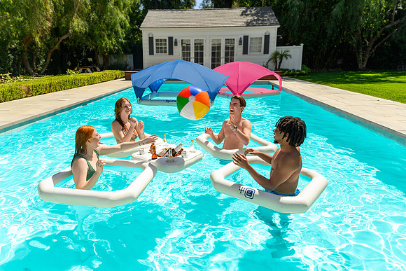 Wide pool lifestyle image of friends relaxing on white floats beneath blue and pink shade canopies.