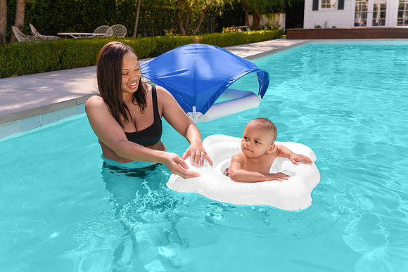 Pool lifestyle photograph of an adult helping a baby seated in a white float in a swimming pool.