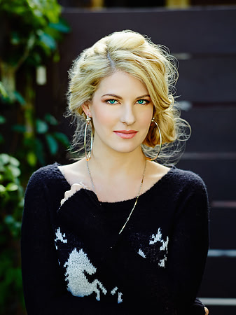 Close editorial portrait of a blonde woman in a black top photographed outdoors with soft greenery.