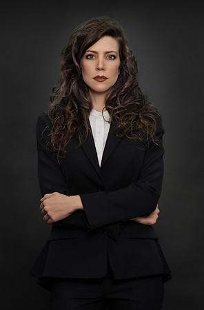 Studio portrait of a brunette woman in a black suit photographed against a charcoal background.