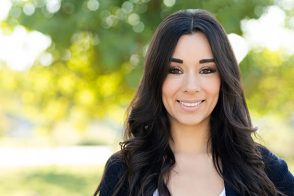 Close professional headshot of a smiling woman with long dark hair against a sunlit outdoor background.