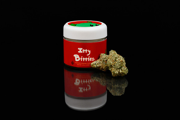 Dark studio product photograph of an Itty Bitties cannabis jar displayed with a cannabis flower bud.