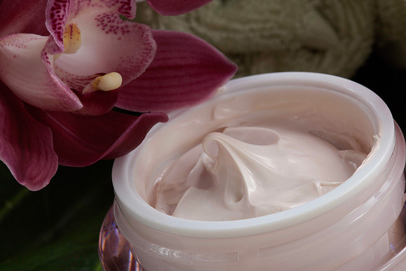 Close beauty product photograph of an open jar of pale pink cream beside a dark pink orchid.