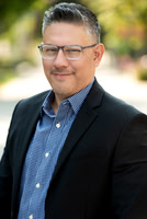 Professional outdoor headshot of a man wearing glasses, a blazer, and blue shirt.