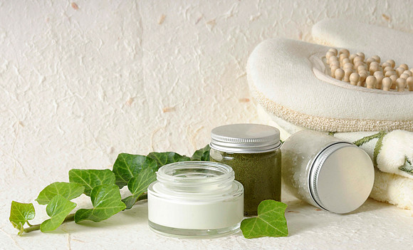 Commercial skincare still life featuring open cosmetic jars, ivy leaves, bath accessories, and silver lids.