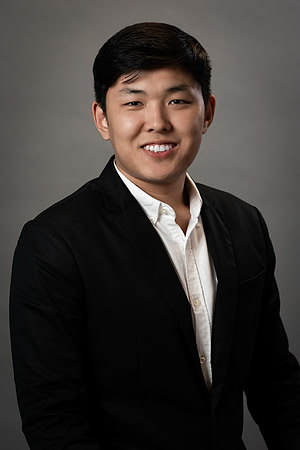 Studio headshot of a smiling man in a black suit against a neutral gray background.