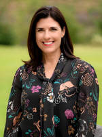 Close business headshot of a smiling woman in a patterned blouse with a softly blurred outdoor background.