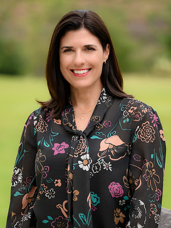 Close business headshot of a smiling woman in a patterned blouse with a softly blurred outdoor background.
