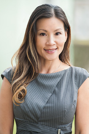 Professional headshot of a woman in a fitted gray dress against a bright office-style background.