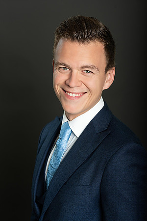 Professional studio headshot of a smiling man in a navy suit and blue tie against dark gray.