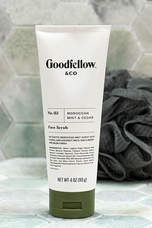 Product photograph of a Goodfellow & Co face scrub tube on pale tile with a dark washcloth in the background.