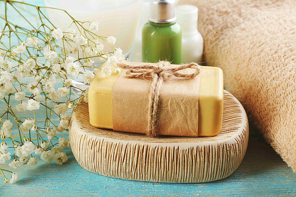 Wellness product still life of a wrapped soap bar on a dish with flowers, towel, and bottles.