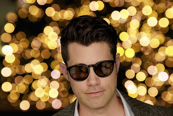 Close editorial portrait of a man in sunglasses against a background of warm circular lights.