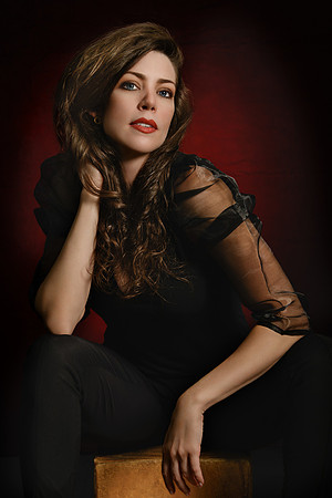 Moody studio portrait of a brunette woman in black against a dark red-and-black background.