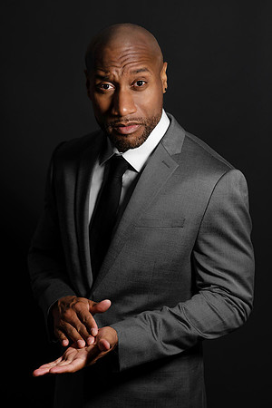Dramatic studio portrait of a man in a gray suit and black tie against a black background.