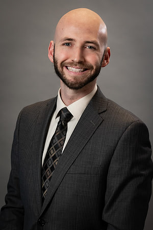 Studio business headshot of a smiling bald man in a dark suit and patterned tie.