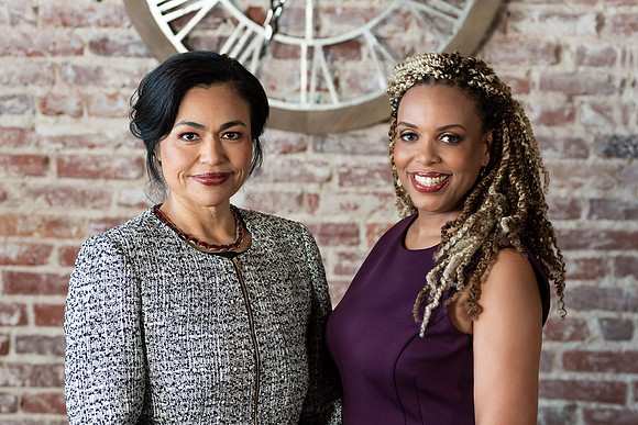 Professional portrait of two women standing together in front of a brick wall.