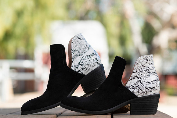 Commercial footwear photograph of black ankle boots with light patterned heel panels styled outdoors.