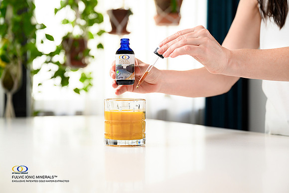 Lifestyle product photograph of a person dispensing Fulvic Ionic Minerals from a dropper into a glass beverage.
