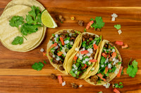 Overhead commercial food photograph of three tacos topped with onion, tomato, cilantro, and lime.