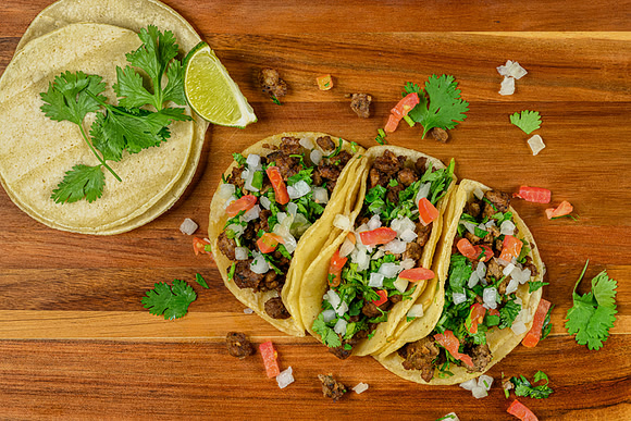 Overhead commercial food photograph of three tacos topped with onion, tomato, cilantro, and lime.