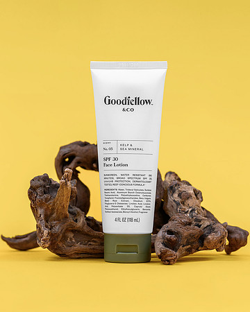 Commercial product photograph of Goodfellow & Co SPF 30 face lotion posed with natural wood pieces on a yellow background.