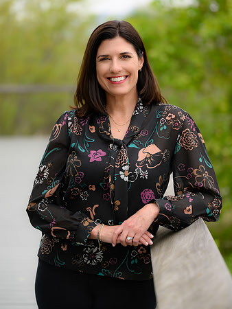 Professional outdoor portrait of a woman in a patterned blouse standing near greenery.