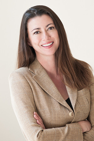 Professional headshot of a woman in a tan blazer with arms crossed against a light background.
