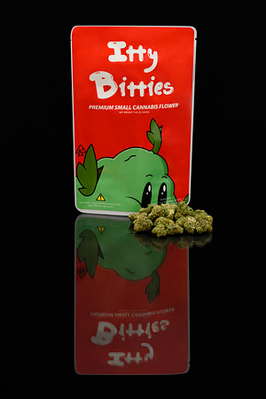 Dark studio product photograph of an Itty Bitties small cannabis flower package with loose buds on a reflective surface.