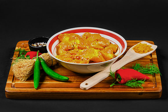 Food product photograph of seasoned raw chicken in a bowl with peppers, herbs, and spices on a wooden board.