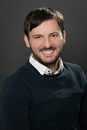 Professional portrait of a smiling man in a dark sweater against a charcoal background.