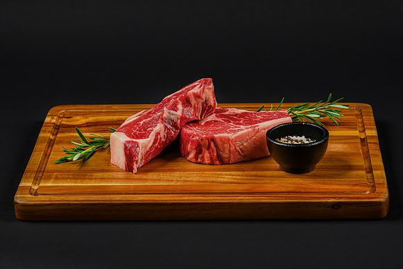 Food product photograph of raw beef steaks on a wooden cutting board with rosemary and seasoning.