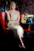 Editorial portrait of a blonde woman seated in a gold evening dress inside a dimly lit interior.