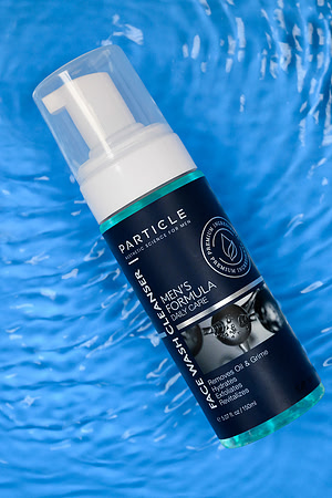 Commercial skincare photograph of a Particle face wash bottle arranged diagonally over a blue water-textured background.