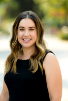 Professional outdoor headshot of a woman in a sleeveless black top with soft greenery behind her.