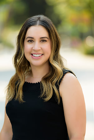 Professional outdoor headshot of a woman in a sleeveless black top with soft greenery behind her.