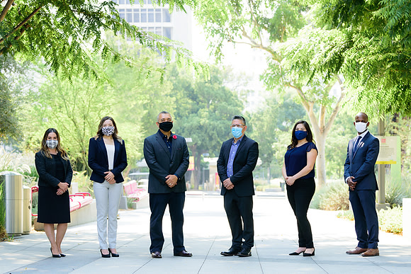 Group business portrait of six professionals standing outdoors while wearing face masks.