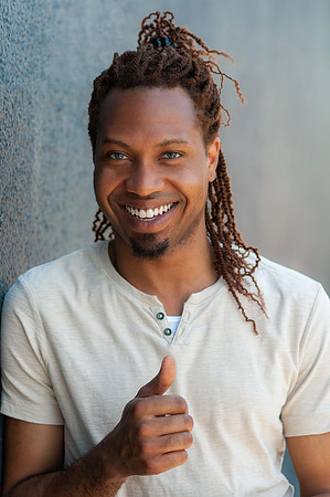 Professional portrait of a smiling man with long braids wearing a light T-shirt against a textured wall.