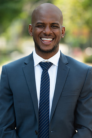 Professional outdoor headshot of a smiling man in a dark suit and striped tie.