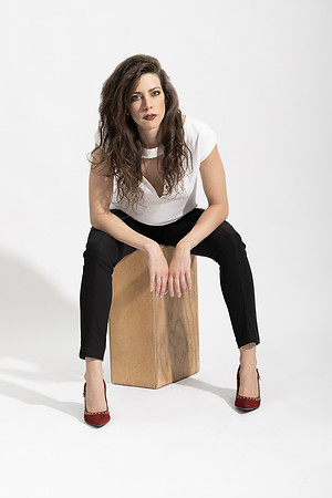 Studio portrait of a brunette woman seated on a wooden block in a white shirt and black pants.