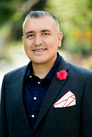 Professional portrait of a man in a dark blazer with a red lapel flower and patterned pocket square.