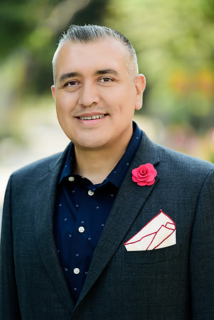 Professional portrait of a man in a dark blazer with a red lapel flower and patterned pocket square.