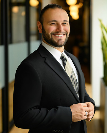Professional portrait of a smiling man in a black suit standing in a modern office corridor.